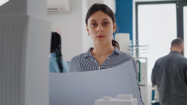 Portrait of architect analyzing blueprints plan to work on construction layout with building model. Woman working on architecture project and urban structure with maquette and print.