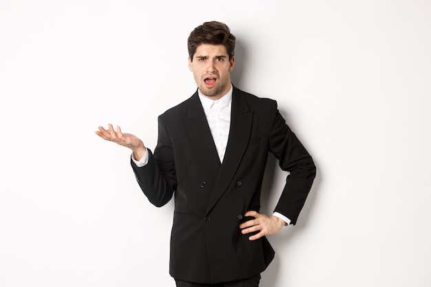 Portrait of arrogant man in black suit, looking confused and disappointed, complaining about something strange, standing over white background