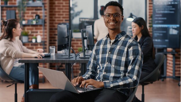 Portrait of artificial intelligence app developer sitting down typing on laptop fixing glasses looking up and smiling at camera. Programer using portable computer innovating cloud computing.