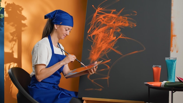 芸術的なツールと創造的なスキルを使用して、キャンバスに水彩絵の具と絵筆で絵を描くアーティストの肖像画。水彩パレットのオレンジ色の絵の具を使用してアートワーク デザインを作成する女性。