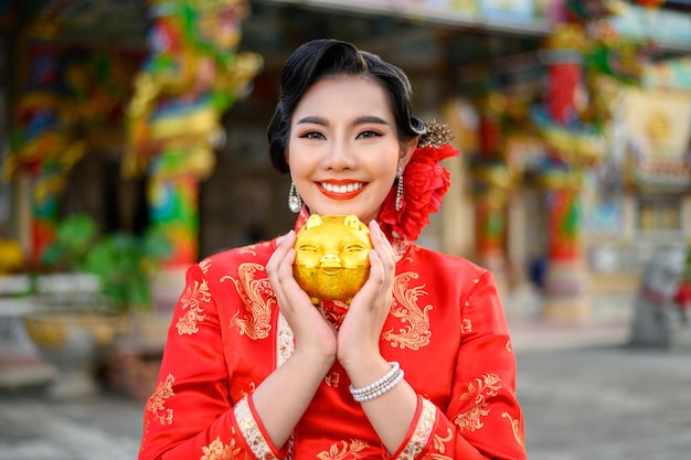 치파오를 입은 아름다운 아시아 여성이 중국 설날 신사에서 황금 돼지 저금통과 함께 웃고 포즈를 취하고 있다