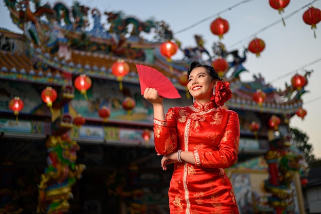 中国の旧正月の神社で笑顔と赤い封筒のファンのポーズを保持しているチャイナドレスを着ている肖像画アジアの美しい女性