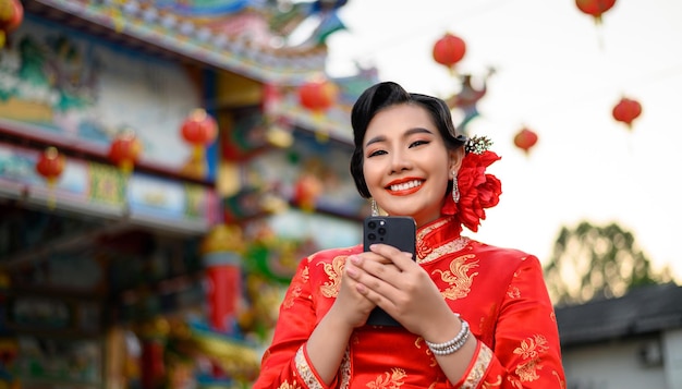 中国の旧正月の神社で笑顔とスマートフォンを使用してチャイナドレスを着ている肖像画アジアの美しい女性