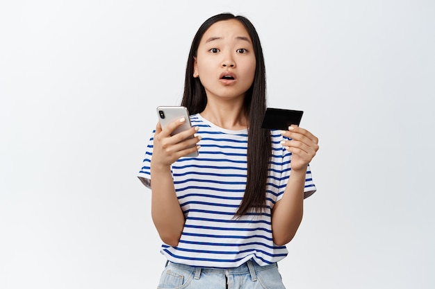 Портрет азиатской брюнетки, выглядящей удивленной, держащей смартфон и кредитную карту, концепция приложения для онлайн-покупок на белом