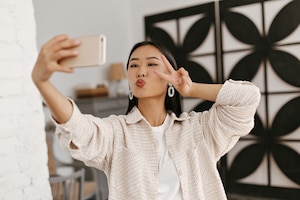Portrait of asian brunette woman in beige jacket takes selfie in cozy room