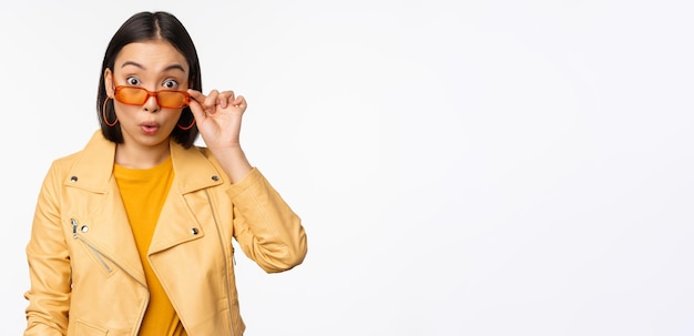 Free photo portrait of asian brunette woman in stylish sunglasses looks surprised and impressed at camera checking out big news wow face expression standing over white background