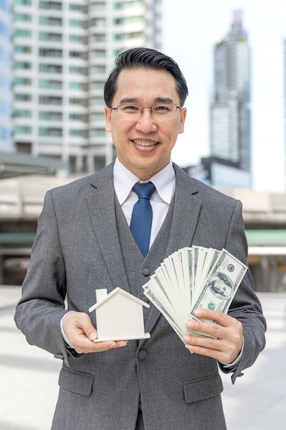 Free photo portrait asian business man holding money us dollar bills and model house on business district