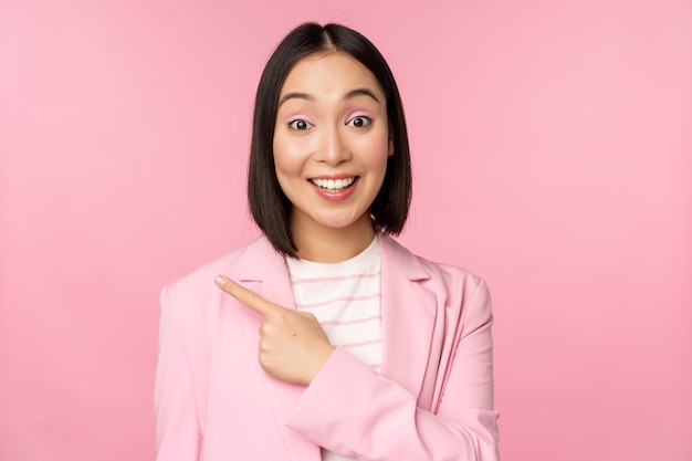 Портрет азиатской деловой женщины-продавщицы в костюме, указывающей пальцем влево, показывая баннерную рекламу, улыбающуюся и выглядящую профессионально на розовом фоне