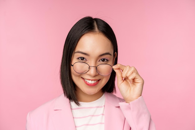 Portrait of asian businesswoman in glasses looking intrigued at camera and smiling professional saleswoman staring with interest pink background