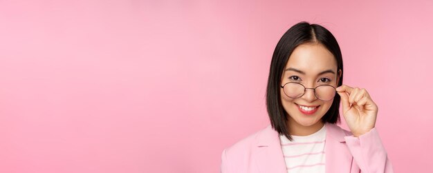 Portrait of asian businesswoman in glasses looking intrigued at camera and smiling professional saleswoman staring with interest pink background