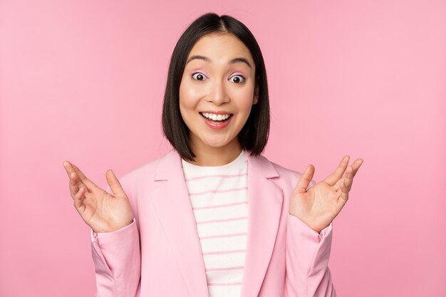 Portrait of asian businesswoman looking surprised at camera clapping hands and staring excited smiling posing against pink background