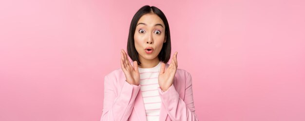 Portrait of asian businesswoman looking surprised at camera clapping hands and staring excited smiling posing against pink background