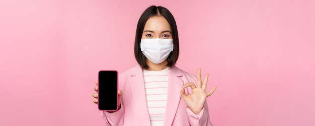 Portrait of asian businesswoman in medical face mask covid19 showing smartphone screen and okay sign pink background