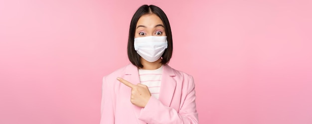 Portrait of asian businesswoman in medical face mask and suit pointing finger left showing advertisement company banner studio pink background