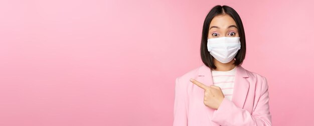 Portrait of asian businesswoman in medical face mask and suit pointing finger left showing advertisement company banner studio pink background