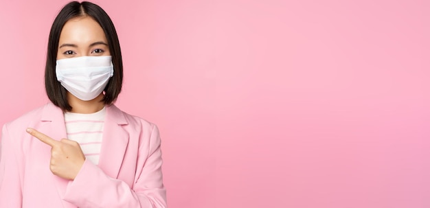 Portrait of asian businesswoman in medical face mask and suit pointing finger left showing advertisement company banner studio pink background