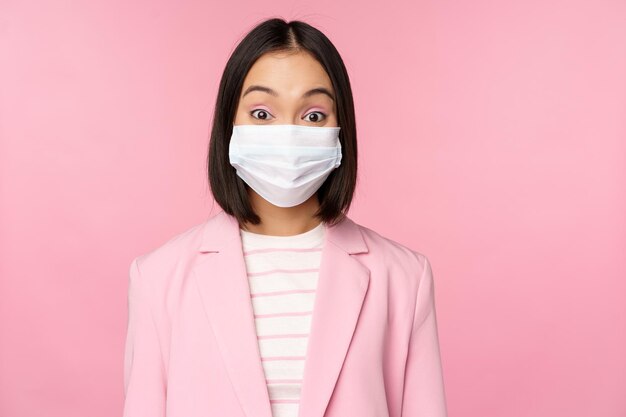 ピンクの背景の上に立っているcovid19パンデミックの間に事務のスーツの概念を身に着けている医療フェイスマスクのアジアの実業家の肖像画