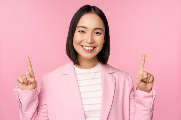 Portrait of asian businesswoman pointing fingers up and smiling showing business company logo information on top standing over pink background