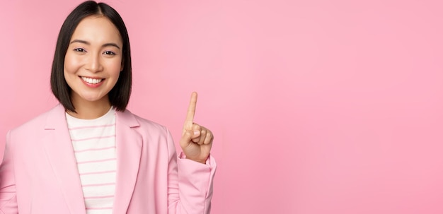 Portrait of asian businesswoman pointing fingers up and smiling showing business company logo information on top standing over pink background