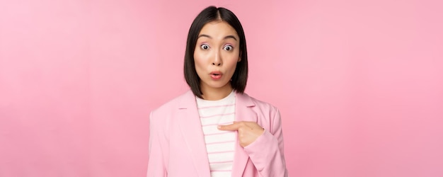 Portrait of asian businesswoman reacts surprised points at herself with disbelief on face posing in suit against pink background