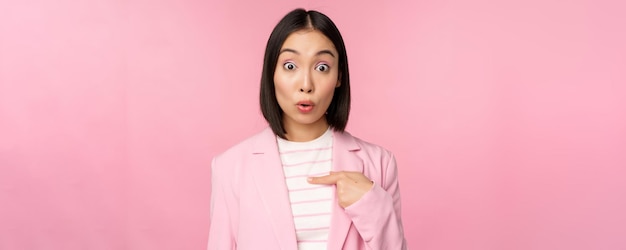 Портрет азиатской деловой женщины удивленно реагирует на себя с недоверием на лице, позируя в костюме на розовом фоне