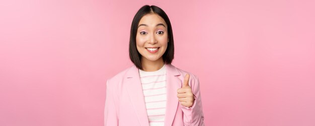Портрет азиатской деловой женщины, довольно улыбающейся, показывающей большие пальцы вверх, хвалит и одобряет стоя в костюме на розовом фоне