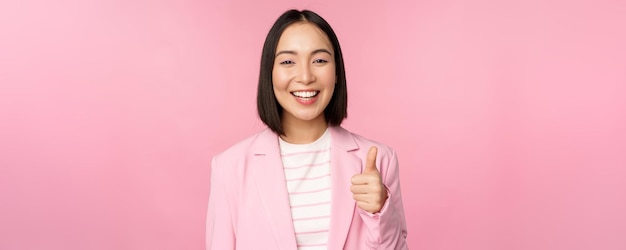 笑顔のアジアの実業家の肖像画は、ピンクの背景の上にスーツを着て立っていることを称賛し、承認するように親指を立てて満足している