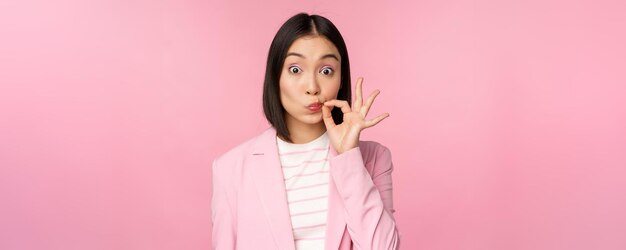 Портрет азиатской корпоративной женщины, показывающей рот, закрывающий губы на ключевом жесте, обещает хранить секрет, стоя на розовом фоне в костюме
