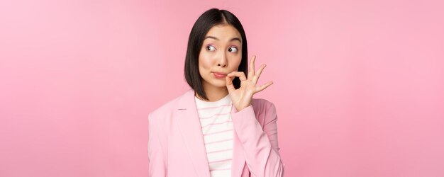 Portrait of asian corporate woman showing mouth seal close shut lips on key gesture promise keep secret standing over pink background in suit