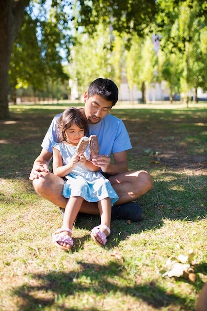 公園でアイスクリームとアジアのお父さんと彼の娘の肖像画。アイスクリームを持って、それを見ている両方の草の上に座っている彼の膝の上に女の子と幸せな男。父性と夏休みの屋外の概念