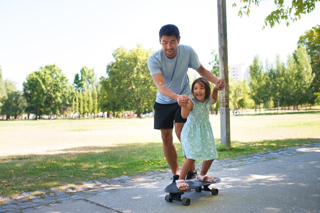 Портрет азиатского папы и маленькой девочки, тренирующихся кататься на скейтборде. Улыбающийся мужчина, идущий по аллее, держит дочь за руки, пока она катается на скейтборде. Активный отдых, здоровый образ жизни и концепция отцовства