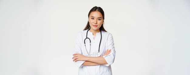 Портрет азиатской женщины-врача, скрещивающей руки, стоящей в медицинской форме и улыбающейся перед камерой со стетоскопом