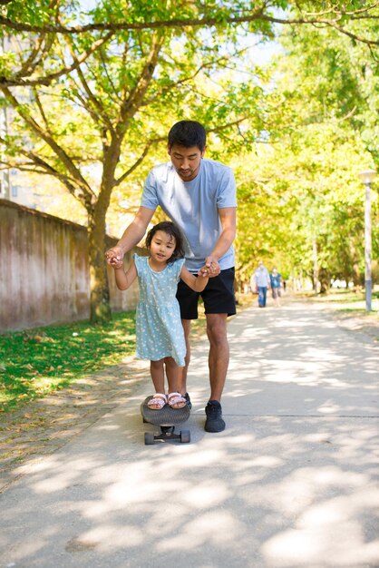 Portrait of Asian father and his daughter skateboarding in park. Happy man holding little girls hands walking on alley while she standing on skateboard. Active rest with kids and fatherhood concept