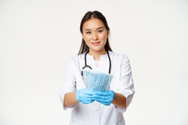 Портрет азиатской женщины-врача, медсестры в резиновых перчатках, дающей стерильные медицинские маски для лица и улыбающейся, меры по предотвращению коронавирусной болезни, белый фон.