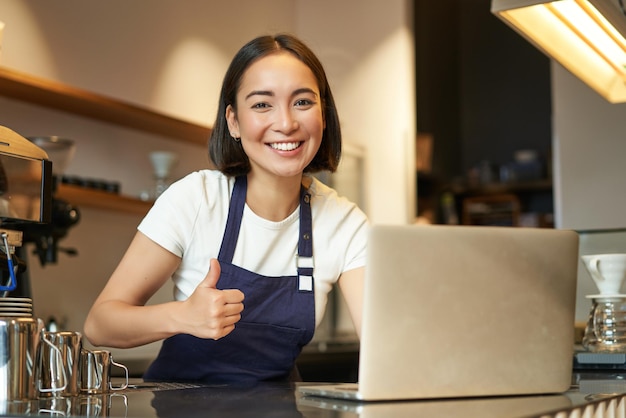 Foto gratuita il ritratto di una ragazza asiatica con il barista in grembiule nella caffetteria mostra i pollici in su, approva o consiglia qualcosa