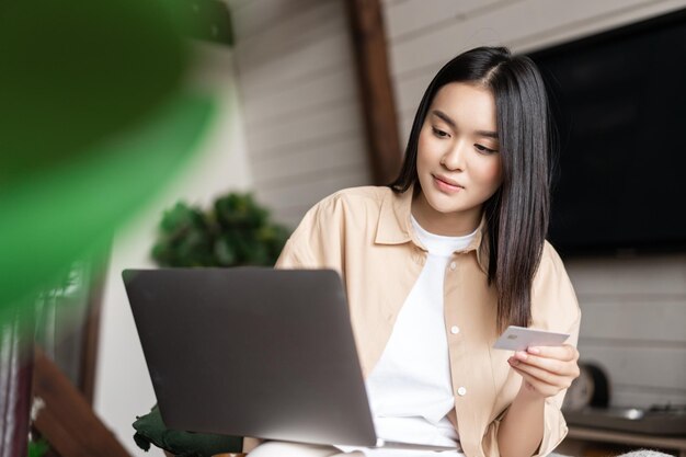 Портрет азиатской девушки, покупающей на веб-сайте с ноутбуком и кредитной картой, молодая женщина покупает онлайн, сидит ...
