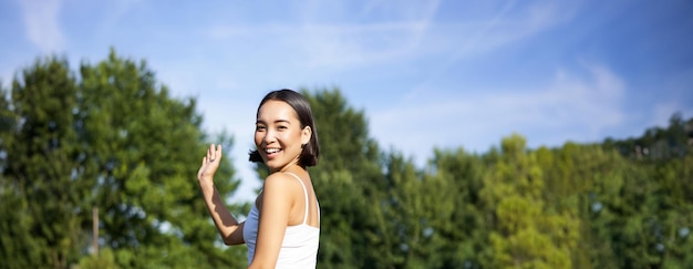 Free photo portrait of asian girl on fitness class in park sitting on rubber mat and wave hand at camera say