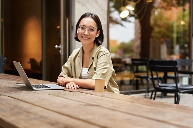 Portrait of asian girl in glasses sitting with laptop in cafe working remotely studying online posin