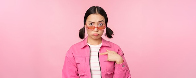 Free photo portrait of asian girl looks confused and points at herself perplexed face stares with disbelief at camera stands over pink background