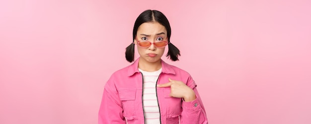 Портрет азиатской девушки выглядит смущенной и указывает на себя озадаченным лицом, недоверчиво смотрит на камеры на розовом фоне