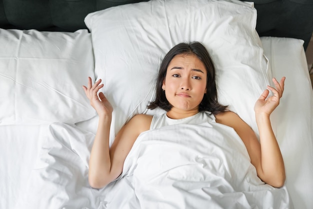 Free photo portrait of asian girl lying in her bed and shrugs looks puzzled overthinking before going to sleep