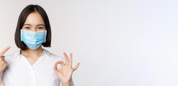 Портрет азиатской девушки в медицинской маске, показывающей знак "хорошо" и указывающей на свою защиту от ковида, стоящую на белом фоне