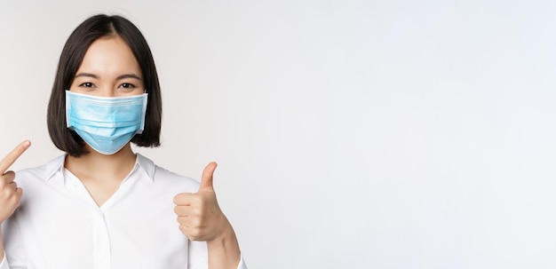 Portrait of asian girl in medical mask showing thumbs up sign and pointing at her covid protection s