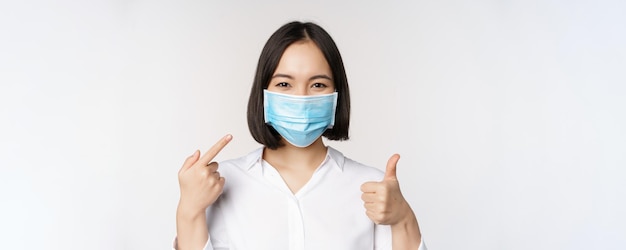 의료용 마스크를 쓴 아시아 소녀 초상화는 엄지손가락을 치켜들고 흰색 배경 위에 서 있는 코비드 보호 장치를 가리키고 있다