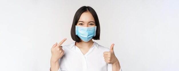 Portrait of asian girl in medical mask showing thumbs up sign and pointing at her covid protection standing over white background