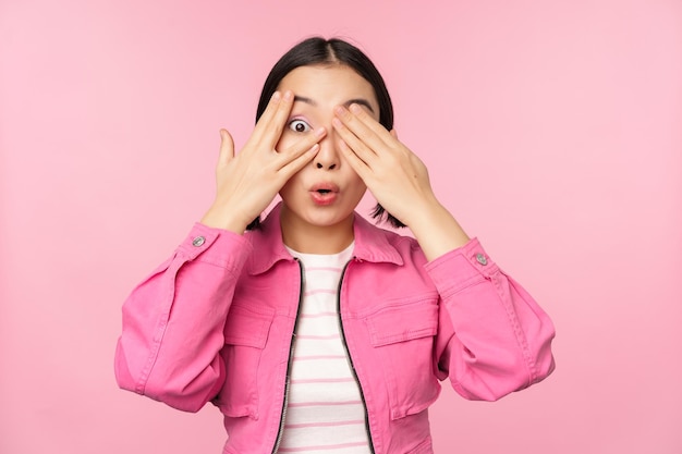 Портрет азиатской девушки с волнением смотрит сквозь пальцы, закрывает глаза, видя удивление, стоящее на розовом фоне
