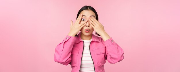 Портрет азиатской девушки с волнением смотрит сквозь пальцы, закрывает глаза, видя удивление, стоящее на розовом фоне