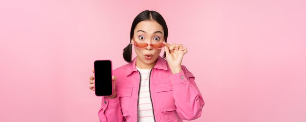 분홍색 배경 복사 공간 위에 놀란 반응을 보이는 휴대폰 화면을 보여주는 아시아 소녀의 초상화