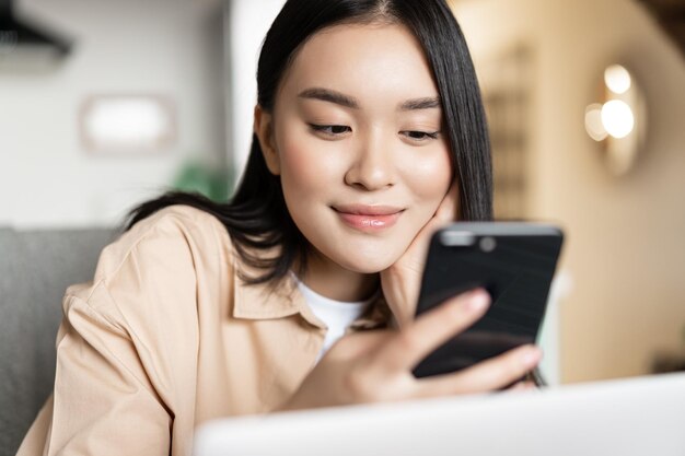 Ritratto di una ragazza asiatica seduta con un laptop che controlla il suo telefono e sorride mentre naviga in siti web su smart...