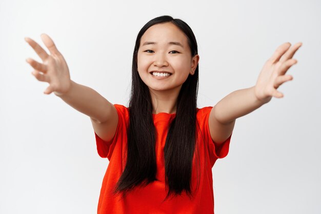 Портрет азиатской девушки, раскинувшей руки для объятий, улыбающейся и выглядящей счастливой, стоящей в красной футболке на белом фоне